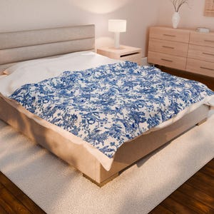 May include: A bed with a beige headboard and a white comforter. A blue and white patterned blanket is draped over the bed. The blanket features a detailed design of birds, flowers, and foliage. The room has a neutral color palette with a wooden floor.