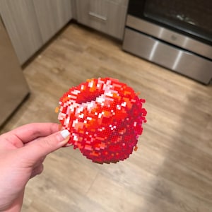 May include: A red and orange pixelated design in the shape of lips. The lips are made of small, square beads, with a white highlight. The item is held in a hand.