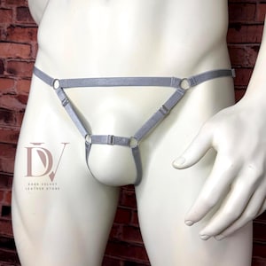 Puede incluir: Un jockstrap de estilo arnés en elástico gris con tiras finas ajustables, anillos metálicos plateados y un deslizador central. El diseño incluye una cintura horizontal conectada a una tira vertical que forma una estructura de bolsa.