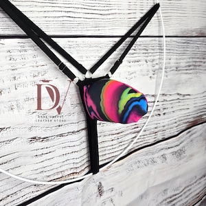 Puede incluir: Un tanga negro con tiras finas ajustables y una bolsa central con un vibrante patrón jaspeado multicolor en rosa neón, amarillo, azul y negro. El logotipo de Dark Velvet Leather Store es visible en el lado izquierdo de la prenda.