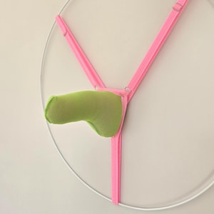 Puede incluir: Una tanga rosa neón con una bolsa verde lima. La prenda está suspendida dentro de un marco circular blanco, creando una composición minimalista y abstracta. Las correas son finas y ajustables.