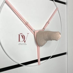 Peut inclure: Une sculpture en cuir rose et beige avec un cadre circulaire. La sculpture présente un harnais rose clair et une forme beige à pois. Le logo "Dark Velvet Leather Store" est visible dans le coin inférieur gauche.