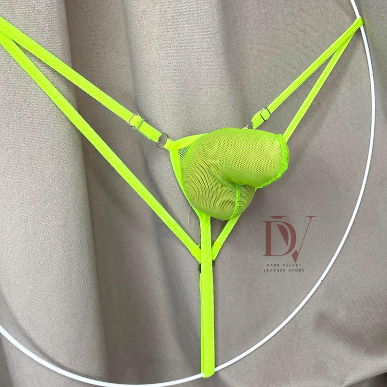 Puede incluir: Tanga de color verde ne&oacute;n brillante con una bolsa de malla y correas ajustables. La lencer&iacute;a est&aacute; suspendida sobre un fondo neutro. El logotipo de Dark Velvet Leather Store es visible en la esquina inferior derecha.
