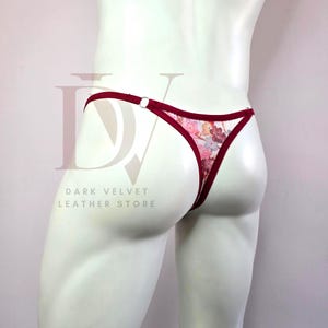 Può includere: Perizoma con stampa floreale con finiture bordeaux e dettaglio con anello argentato. La biancheria intima presenta un delicato motivo floreale nelle tonalità del rosa, viola e bianco. Il testo "DARK VELVET LEATHER STORE" è visibile.