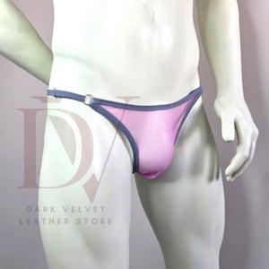 Puede incluir: Tanga rosa con ribete gris y un pequeño detalle de anillo metálico. La ropa interior está hecha de un material transparente. El logotipo de Dark Velvet Leather Store es visible en el fondo.