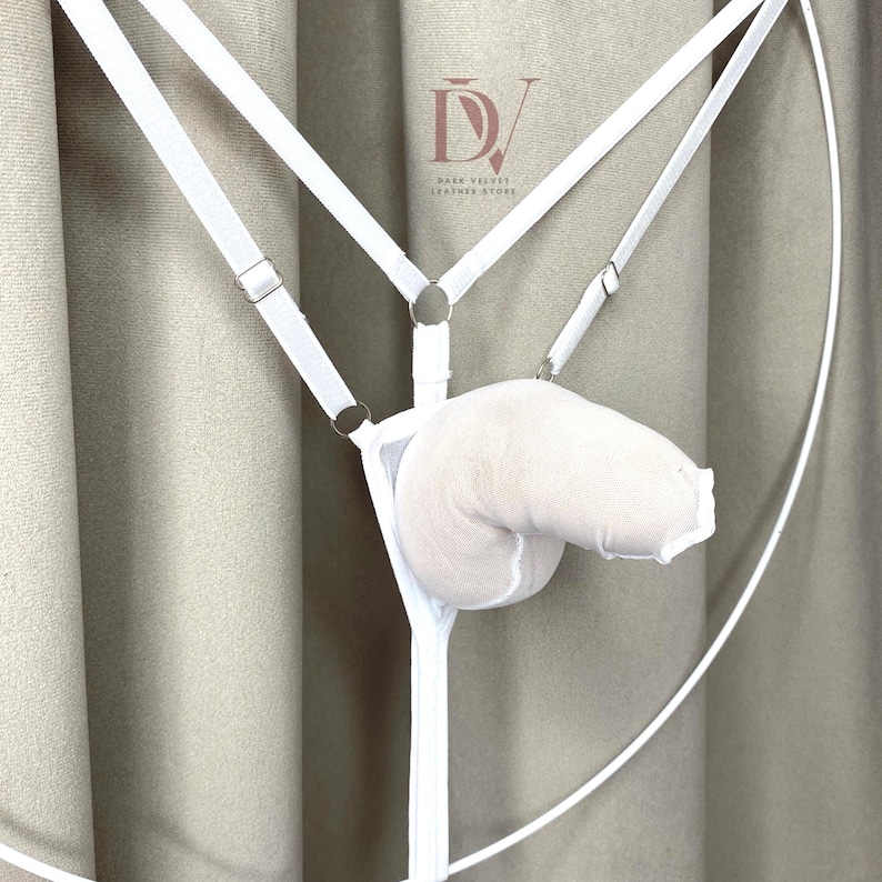 Puede incluir: Conjunto de lencer&iacute;a blanco con dise&ntilde;o de arn&eacute;s, correas ajustables y herrajes met&aacute;licos. Incluye una bolsa suave y acolchada. El logotipo de Dark Velvet Leather Store es visible en el fondo.