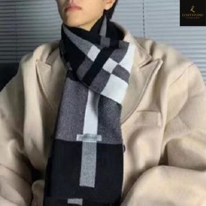 Puede incluir: Una bufanda a cuadros negros, blancos y grises. La bufanda está drapeada alrededor del cuello y el pecho. La bufanda tiene forma rectangular con un patrón de cuadros y un borde negro liso. El fondo es un abrigo beige.