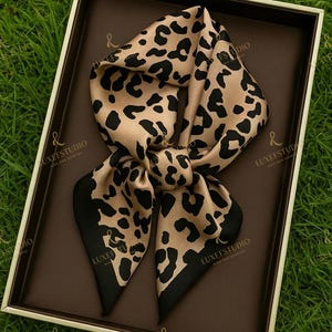 May include: A tan and black leopard print scarf, knotted in the center, rests in a brown box with a white border. The scarf has black borders and the text "LUXEFSTUDIO" is visible on the box.