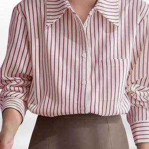 Puede incluir: Una camisa blanca con rayas verticales rojas. Tiene cuello, mangas largas y un bolsillo en el pecho. La camisa está abotonada y se combina con una falda marrón.