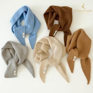 Puede incluir: Cinco bufandas de punto suave en varios colores: azul claro, marrón, beige, gris y camel. Cada bufanda tiene una etiqueta rectangular y un diseño anudado. Las bufandas están dispuestas sobre una superficie blanca.