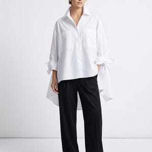 Könnte beinhalten: Ein weißes, langärmliges Hemd mit zwei Vordertaschen und einem High-Low-Saum, kombiniert mit einer schwarzen Hose mit weitem Bein. Das Hemd hat Bindedetails an den Manschetten. Das Outfit ist ein klassischer, minimalistischer Stil.