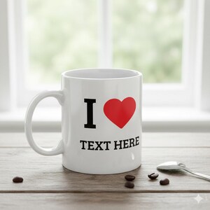 May include: White ceramic mug featuring a black "I" and a red heart design, with the words "TEXT HERE" in black. The mug is on a wooden surface with coffee beans and a spoon.