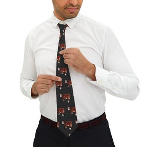 Puede incluir: Una corbata negra con un patrón repetido de vacas marrones y blancas. La corbata es ajustada por una persona que lleva una camisa blanca y pantalones negros. La corbata es de ancho estándar y está hecha de tela lisa.