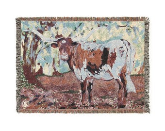 Op de afbeelding: Geweven sprei met een Longhorn-koe in een veld. De koe heeft bruine en witte vlekken met lange hoorns. De achtergrond bevat bomen en bladeren in groen, bruin en blauw. De deken heeft een gefranjerde rand.