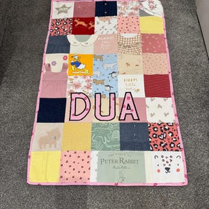 May include: A patchwork baby quilt with a pink border. The quilt is made of various fabric squares, including prints with animals, floral patterns, and the name "DUA" in large, pink letters. The quilt measures approximately 91 x 122 cm.