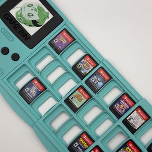 May include: A teal game cartridge holder shaped like a retro handheld console, labeled "GAME BIN" and "NIATO". It holds multiple Nintendo Switch game cartridges, with game titles visible, including "Just Dance 2022" and "Crash Bandicoot".