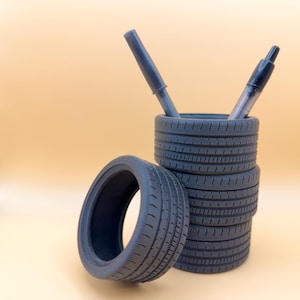 May include: A unique desk organizer made from stacked miniature tires. The gray tires form a pen holder, with two pens inside. One tire is placed separately. The background is a solid light orange.