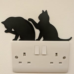 May include: A black silhouette of two cats positioned above a white double light switch plate. One cat is sitting and grooming itself while the other cat stands beside it with one paw raised. The cats are flat, matte, and cut into a cat shape.