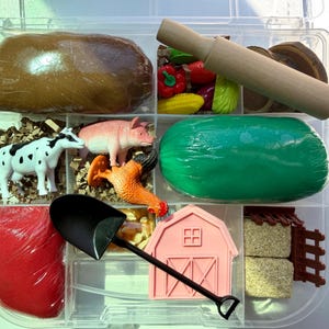May include: A clear plastic container with various farm-themed toys. Includes a wooden rolling pin, toy vegetables, a cow, pig, and chicken figurines, a pink barn, a black shovel, and wrapped play dough in brown, red, and green.