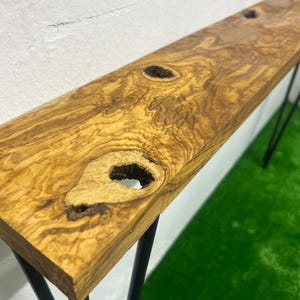 May include: A long, narrow wooden console table with a natural, olive-toned finish. The tabletop features unique knot details and is supported by black metal hairpin legs. The table is approximately 60 inches long.