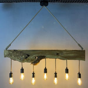 Könnte beinhalten: Ein rustikaler Holz-Kronleuchter mit acht Edison-Glühbirnen. Die Leuchte wird mit Seilen an einer schwarzen Deckenhalterung aufgehängt. Der Holzbalken hat ein natürliches, unbehandeltes Aussehen.