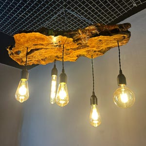 Puede incluir: Una lámpara colgante con una gran losa de madera de borde natural como base. Cinco cables negros suspenden cinco bombillas de cristal estilo Edison de diferentes formas y tamaños, emitiendo un brillo cálido y brillante desde la madera.