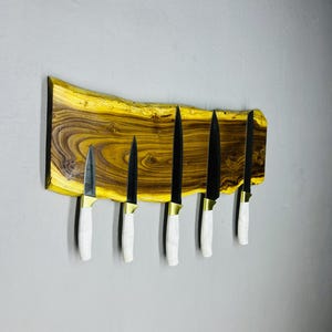 May include: A wooden knife holder with six knives. The holder is made of a live edge wood with a yellow and brown grain. The knives have black blades, white handles, and gold-colored bolsters. The knives are mounted on the wooden holder.