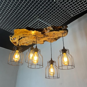 May include: A rustic, wooden beam chandelier with five Edison-style light bulbs encased in black metal cages. The wooden beam has a natural, golden-brown color and is suspended from a black grid ceiling.