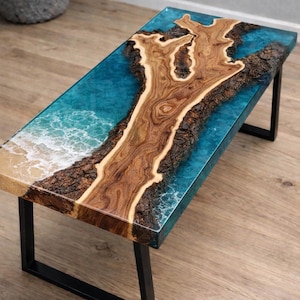 May include: A rectangular coffee table with a unique design. The tabletop features a wooden centerpiece surrounded by a blue resin, resembling an ocean scene. The table is supported by black metal legs.