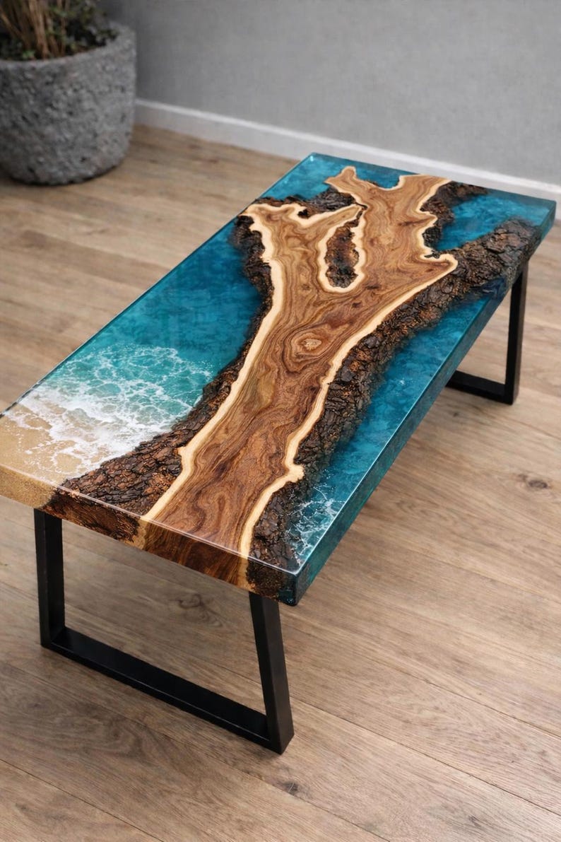 May include: A rectangular coffee table with a unique design. The tabletop features a wooden centerpiece surrounded by a blue resin, resembling an ocean scene. The table is supported by black metal legs.