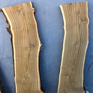 May include: Two unfinished, light brown wood slabs with natural edges. The wood grain is visible, and the edges have a darker brown bark. The slabs are rectangular with irregular, natural edges, suitable for woodworking or rustic decor.