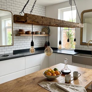May include: A rustic wooden beam pendant light fixture with five hanging Edison bulbs suspended by black chains above a kitchen island. The kitchen features white subway tile backsplashes, black worktops, a farmhouse sink, and two gold-framed mirrors.