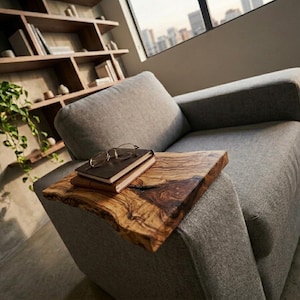 Op de afbeelding: Een grijze gestoffeerde fauteuil met een bijzettafel van natuurlijk hout. De bijzettafel heeft een boomstamrand en bevat een stapel boeken en een bril. Een boekenplank en een raam zijn op de achtergrond.