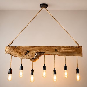 May include: A linear pendant light fixture featuring a natural live-edge wooden beam suspended by thick jute rope. Seven black sockets hang from the beam, each fitted with a clear Edison-style light bulb, creating a warm, downward-facing glow.