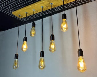 May include: A handmade light fitting with a golden-yellow wooden beam and seven Edison-style light bulbs suspended by black cords. The bulbs are illuminated, emitting a warm light. The fixture is mounted on a black grid ceiling.