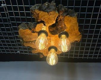 May include: A ceiling light fixture featuring a natural, irregular wood slab base with a live edge. Three black metal sockets are mounted to the wood, each holding a clear glass Edison-style light bulb with visible vertical filaments.