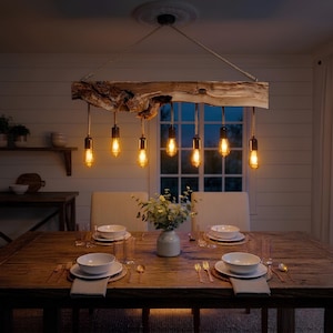 May include: A wooden chandelier with six Edison-style light bulbs hangs above a wooden dining table. The light fixture is suspended by a rope. The dining table is set with white plates, cutlery, and napkins. The room has a warm, inviting ambiance.