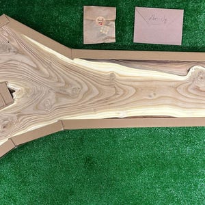 May include: A large, light-colored wooden slab with a natural edge, packaged in a custom cardboard box. The wood grain is prominent, showcasing various shades of brown and tan. Two small paper items are visible nearby on a green surface.