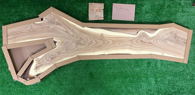 May include: A large, light-colored wooden slab with a natural edge, packaged in a custom cardboard box. The wood grain is prominent, showcasing various shades of brown and tan. Two small paper items are visible nearby on a green surface.