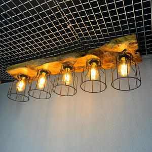 May include: A wooden light fitting featuring five Edison-style bulbs within black wire cages. The fixture is mounted on a natural-edge wooden beam, creating a warm, industrial aesthetic. The bulbs are lit, casting a warm glow. The beam is approximately 120 cm long.