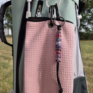 Puede incluir: Una toalla de golf rosa claro colgada de una bolsa de golf verde menta. Un dije de cuentas con cuentas moradas, rosas y plateadas está sujeto a la toalla con un mosquetón negro. La toalla tiene un tejido de gofre texturizado.