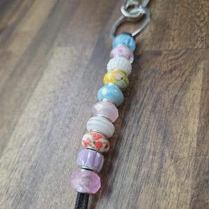 May include: A beaded keychain featuring a variety of colorful beads in shades of pink, blue, white, and yellow. The beads are strung on a black cord and attached to a silver-colored clasp. The beads have different patterns and textures.