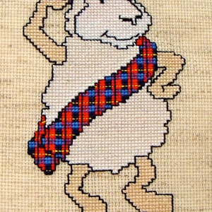 May include: A white sheep wearing a black and red plaid kilt and a black hat with a red pom-pom. The sheep is standing on its hind legs and has its front legs raised in the air. The sheep is smiling and has a playful expression on its face. The image is a cross-stitch design.