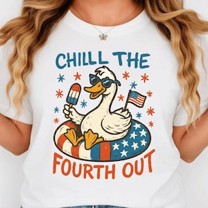 Può includere: T-shirt bianca con una grafica di un'anatra dei cartoni animati con occhiali da sole che galleggia su un salvagente. L'anatra tiene in mano un ghiacciolo e una piccola bandiera americana. Il testo recita "CHILLL THE FOURTH OUT."