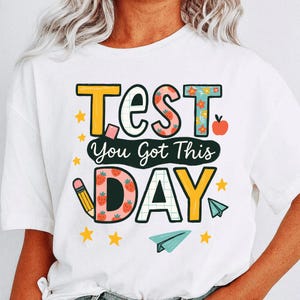 May include: White t-shirt with a colorful graphic design. The design includes the word "TEST" in yellow and strawberry-patterned letters, along with the phrase "You Got This DAY". A pencil, stars, apple, and paper airplane complete the design.