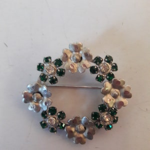 May include: A silver brooch with a circular design of green and clear rhinestones set in a flower shape.