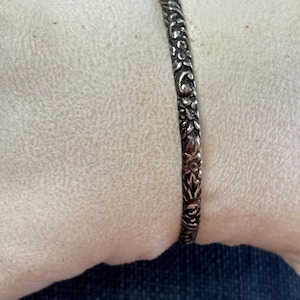 May include: A silver-toned metal bangle bracelet featuring an intricate, embossed floral pattern that covers the entire outer surface. The design consists of repeating flower motifs and leafy vines with darkened recessed areas to highlight the details.