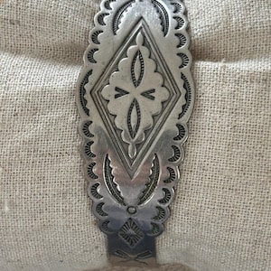 May include: A silver cuff bracelet featuring an elongated diamond-shaped center panel with intricate stamped detailing, scalloped edges, and linear engravings. The metal surface displays a polished finish with darkened recessed areas.