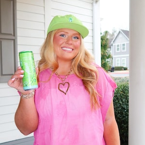 May include: A woman wearing a pink top, white shorts, and a green cap holds a green beverage can. The can has a white and green label. She is wearing gold bracelets and a gold heart pendant necklace. The word "AFTER" is above her head.