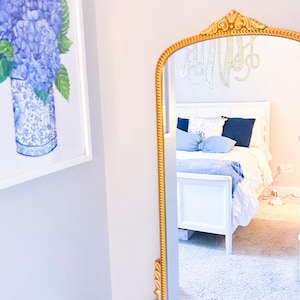 以下が含まれることがあります： 白いベッド、青いクッション、金色のフレームの鏡がある寝室のシーン。花瓶に入った青いアジサイの水彩画が壁に掛かっています。鏡はベッドを映し出し、「AFTER:」の文字が見えます。